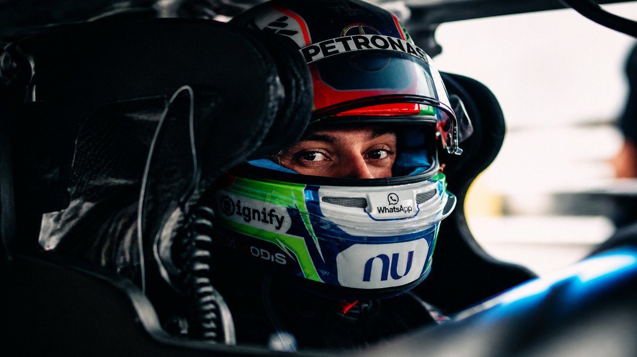 Close-up of a race car driver wearing a helmet with PETRONAS branding, sitting in the car cockpit.